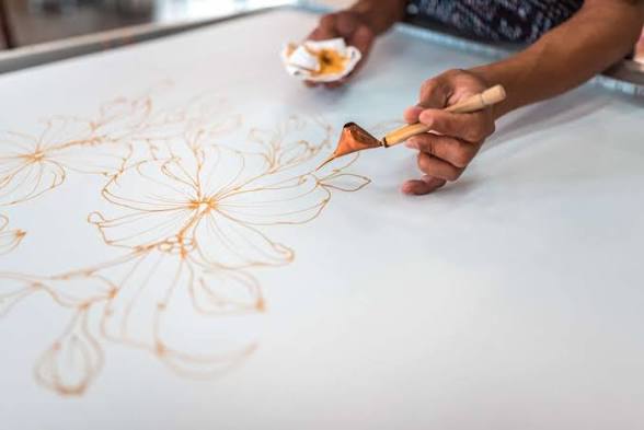 Close-up of a batik artisan drawing floral wax outlines on cloth with a traditional canting tool